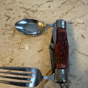 May include: A stainless steel camping utensil with a spoon, fork, and knife. The handle is a reddish-brown color. The utensil is designed to fold up for easy storage and transport.