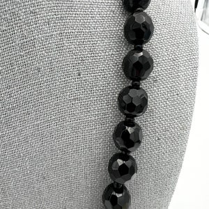 May include: A close-up of a black beaded necklace. The beads are large, faceted, and have a glossy finish. The necklace is displayed against a gray fabric background.