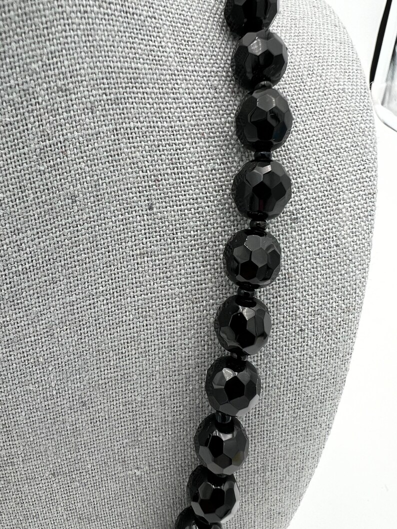 May include: A close-up of a black beaded necklace. The beads are large, faceted, and have a glossy finish. The necklace is displayed against a gray fabric background.