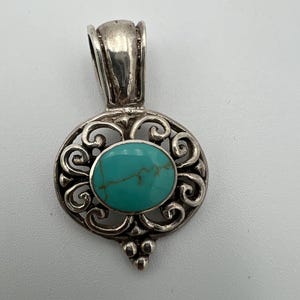 May include: A silver pendant with an oval turquoise stone set in an ornate, openwork design. The pendant has a bail at the top for a chain and a decorative bottom. The turquoise stone is a vibrant blue-green color.