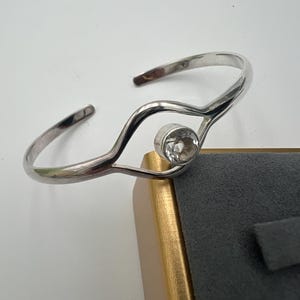 Puede incluir: Un brazalete plateado con una piedra preciosa redonda y transparente en el centro. El brazalete tiene un diseño abierto y fluido. La piedra está engastada en un bisel. El brazalete se exhibe en una caja de joyería gris y dorada.