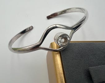 May include: A silver-toned cuff bracelet with a clear, round gemstone set in the center. The bracelet has a flowing, open design. The gemstone is set in a bezel setting. The bracelet is displayed on a gray and gold jewelry box.