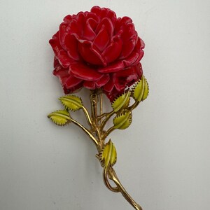 May include: A red rose brooch with a detailed, carved design. The rose is the focal point, with a gold-tone stem and green and yellow leaves. The brooch is a classic floral accessory.