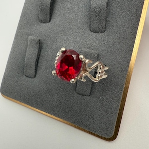 May include: A silver ring with a large, oval-cut, bright red gemstone. The ring has an ornate, openwork design on the sides. The ring is displayed on a gray velvet jewelry display.