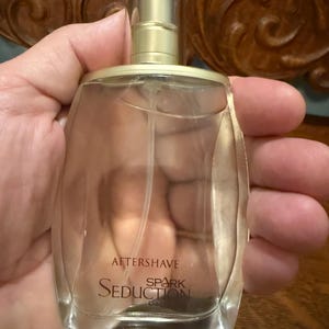 May include: A clear glass bottle of aftershave with a gold-colored cap and a black top. The bottle has the words "AFTERSHAVE", "Seduction", and "Spark" printed on it. The bottle is held in a hand.