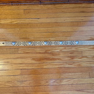 May include: A light-colored belt with heart-shaped embellishments. The belt features a heart-shaped buckle and several heart-shaped studs along its length. The belt is lying on a wooden surface.