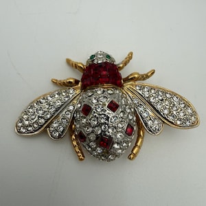 May include: A gold-toned bee brooch with rhinestone and red jewel accents. The brooch features a detailed design with clear rhinestones on the wings and body, and red jewels on the head and body. The bee has gold-colored legs and antennae.