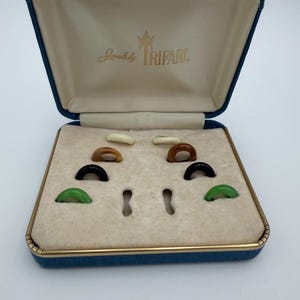 May include: A blue jewellery box with gold trim and the text "Jewels by Trifari" contains several small, colourful rings. The rings are in shades of green, black, brown, and cream. The box is open, revealing a cream interior.