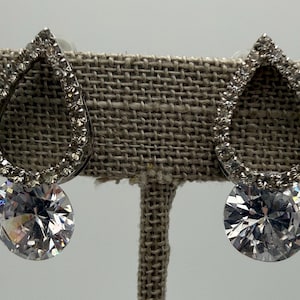 May include: A pair of silver-toned earrings featuring a teardrop-shaped frame encrusted with small clear stones. Each earring has a large, round, faceted clear stone dangling from the bottom of the teardrop frame.