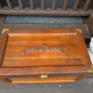Puede incluir: Un cofre rectangular de madera con un asa decorativa tallada y esquinas de lat&oacute;n. El cofre est&aacute; hecho de madera te&ntilde;ida y tiene un pestillo de lat&oacute;n. El cofre mide aproximadamente 61 cm de largo, 36 cm de ancho y 30 cm de alto.