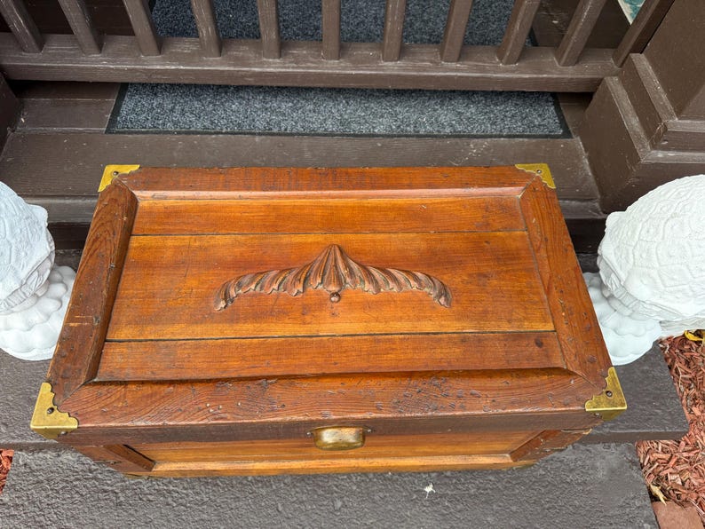 Puede incluir: Un cofre rectangular de madera con un asa decorativa tallada y esquinas de lat&oacute;n. El cofre est&aacute; hecho de madera te&ntilde;ida y tiene un pestillo de lat&oacute;n. El cofre mide aproximadamente 61 cm de largo, 36 cm de ancho y 30 cm de alto.