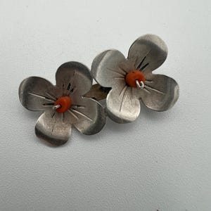 May include: A pair of silver-toned flower-shaped earrings. Each earring features three petals and a small, orange bead in the center. The earrings are designed to resemble a flower. The petals have etched lines for detail.
