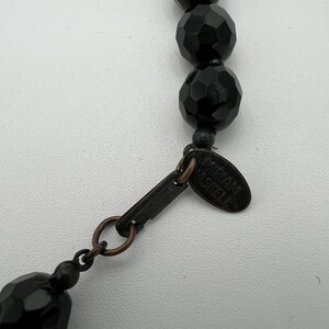 May include: Close-up of a black beaded necklace with faceted beads. The necklace has a metal clasp and a tag that reads "Miriam Haskell". The beads are a deep black color and have a glossy finish. The necklace is on a white background.