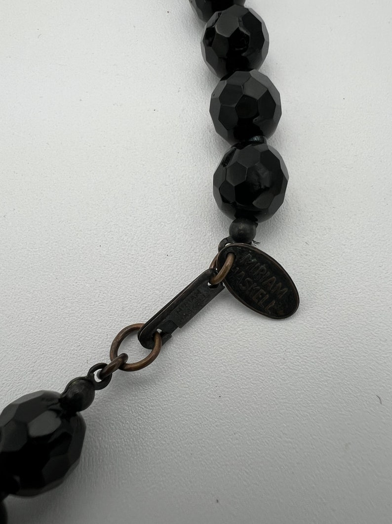 May include: Close-up of a black beaded necklace with faceted beads. The necklace has a metal clasp and a tag that reads "Miriam Haskell". The beads are a deep black color and have a glossy finish. The necklace is on a white background.