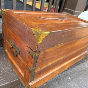 Puede incluir: Un ba&uacute;l de madera vintage con protectores de esquina y herrajes de lat&oacute;n. El ba&uacute;l es de color marr&oacute;n c&aacute;lido, con un dise&ntilde;o tallado en la parte superior. Tiene un asa met&aacute;lica en el lateral. El ba&uacute;l probablemente se utiliza para almacenamiento o decoraci&oacute;n.