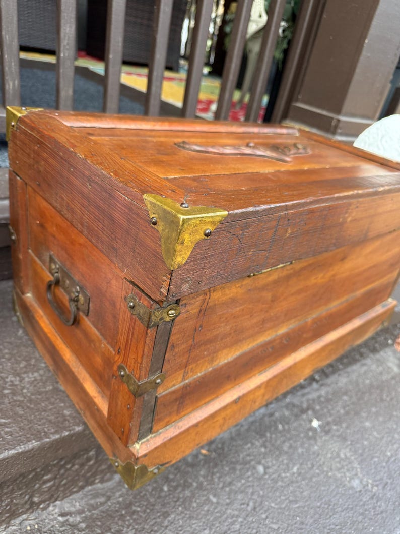 Puede incluir: Un ba&uacute;l de madera vintage con protectores de esquina y herrajes de lat&oacute;n. El ba&uacute;l es de color marr&oacute;n c&aacute;lido, con un dise&ntilde;o tallado en la parte superior. Tiene un asa met&aacute;lica en el lateral. El ba&uacute;l probablemente se utiliza para almacenamiento o decoraci&oacute;n.