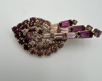 May include: Vintage brooch featuring a cluster of purple and pink rhinestones in various shapes and sizes. The brooch has a gold-tone metal setting and a unique, asymmetrical design. A classic piece of jewelry.