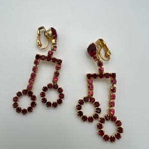 May include: A pair of vintage clip-on earrings featuring a musical note design. The earrings are adorned with red and pink rhinestones set in a gold-tone metal. The circular base of the note is a darker red, while the note's stem is a lighter pink.