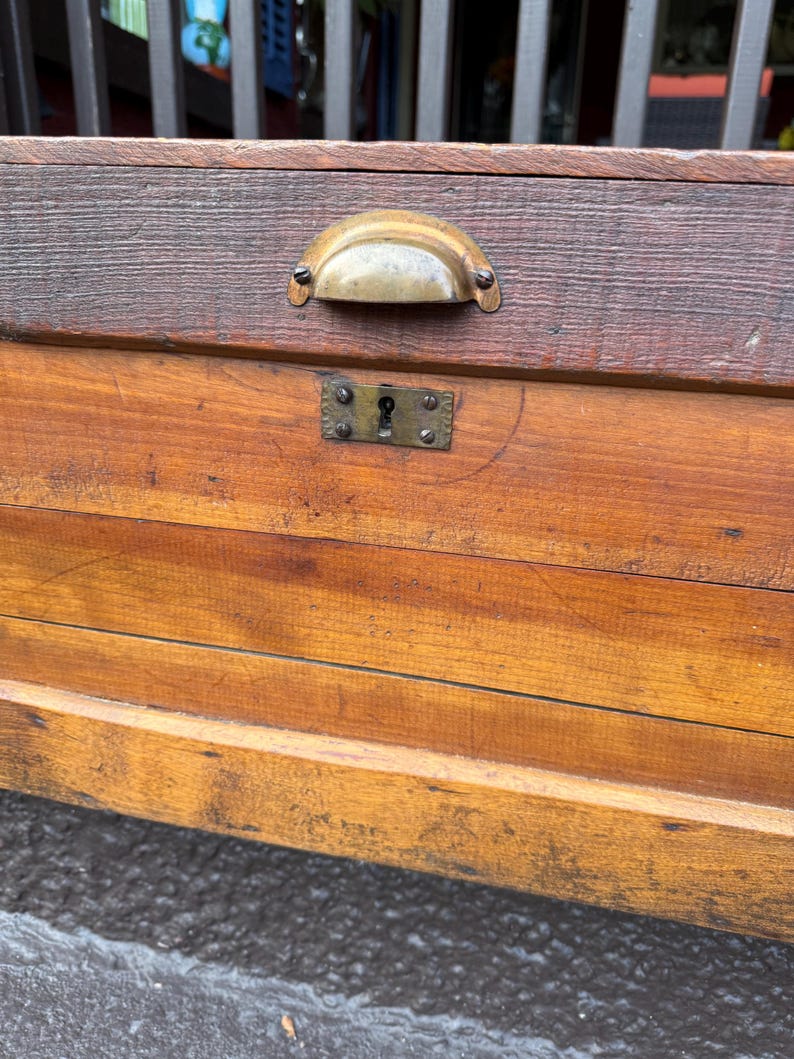 Puede incluir: Primer plano de un cofre de madera vintage con un asa y cerradura de lat&oacute;n. El cofre est&aacute; hecho de madera te&ntilde;ida con vetas visibles y una parte superior oscura. La ferreter&iacute;a tiene un aspecto deslustrado.