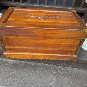 Puede incluir: Un cofre de madera rectangular con un acabado marr&oacute;n c&aacute;lido. El cofre presenta protectores de esquina de lat&oacute;n y detalles decorativos. La tapa tiene un dise&ntilde;o tallado y el cofre parece ser vintage o antiguo.
