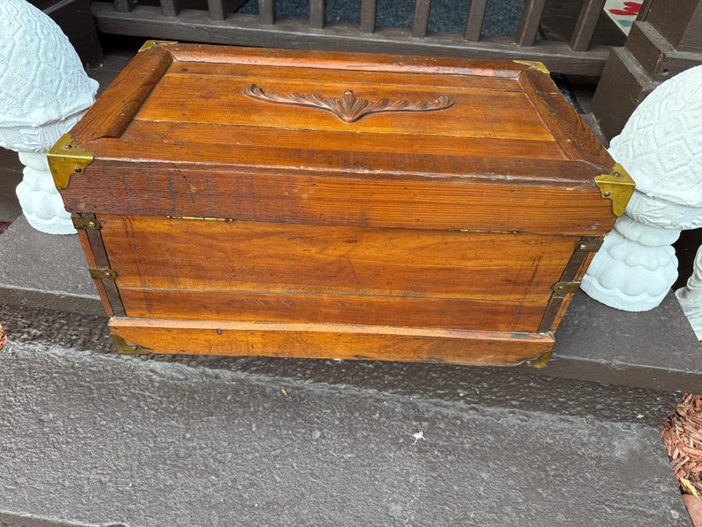 Puede incluir: Un cofre de madera rectangular con un acabado marr&oacute;n c&aacute;lido. El cofre presenta protectores de esquina de lat&oacute;n y detalles decorativos. La tapa tiene un dise&ntilde;o tallado y el cofre parece ser vintage o antiguo.