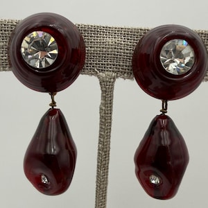 May include: A pair of vintage clip-on earrings. Each earring features a dark red, circular top with a clear, sparkling rhinestone center. A teardrop-shaped, dark red pendant hangs below, also with a rhinestone accent. The earrings are attached with gold-toned metal.