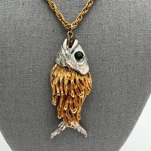 May include: A gold-toned chain necklace with a fish pendant. The fish pendant has a silver-colored head with a green gem eye and a gold-colored body. The fish's tail is silver-colored.