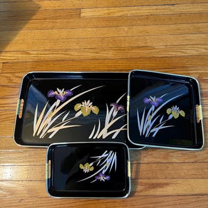 May include: Set of three black serving trays with a floral design. The trays feature a gold trim and woven handles. The floral design includes purple, yellow, and white iris flowers and blades of grass.