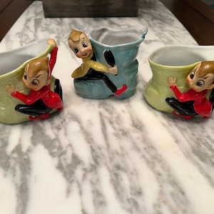 Puede incluir: Tres macetas de cerámica vintage con forma de bota, cada una con una figura de elfo caprichosa trepando por el lateral. Las botas son en tonos verdes y azules, con los elfos vestidos con trajes rojos y negros. Un objeto decorativo encantador.