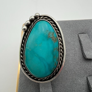 Op de afbeelding: Ovale turkoois ring met een zilveren zetting. De turkoois steen is een levendige blauwgroene kleur met bruine aders. De ring heeft een gevlochten zilveren rand en kleine zilveren kralen aan de bovenkant.