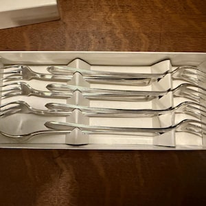 May include: A set of six silver forks arranged in a white box. The forks have a classic design with three tines and a curved handle. The box is open, revealing the forks neatly placed within.