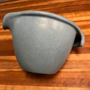 May include: A speckled, light blue ceramic mixing bowl with a pouring spout. The bowl has a rounded shape with a curved rim and a handle-like extension on one side. The bowl is sitting on a wooden surface.