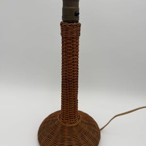 May include: A table lamp base featuring a tall, cylindrical stem and a wide, circular, domed base, both constructed from woven brown wicker. The top of the lamp includes a dark metal socket with a small black switch and a light-colored power cord.