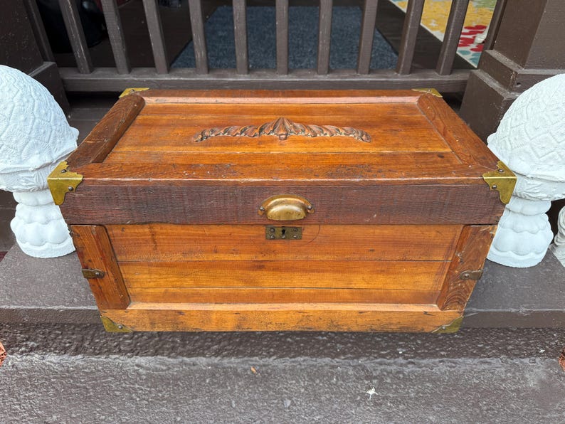 Puede incluir: Un ba&uacute;l de madera vintage con herrajes de lat&oacute;n. El ba&uacute;l tiene forma rectangular con un dise&ntilde;o tallado en la tapa. La madera tiene un tono marr&oacute;n rojizo c&aacute;lido. El ba&uacute;l mide aproximadamente 76 cm de ancho, 46 cm de profundidad y 51 cm de alto.
