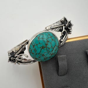 May include: A silver bracelet featuring a large, oval turquoise stone with brown veining. The bracelet has ornate floral and leaf detailing on the sides, with a polished finish. The turquoise stone is set in a silver bezel.