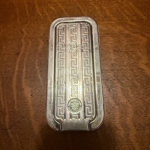May include: A rectangular, silver-toned metal object with a Greek key design. The object has a slightly curved top and a small, circular emblem at the bottom. The surface shows signs of wear.