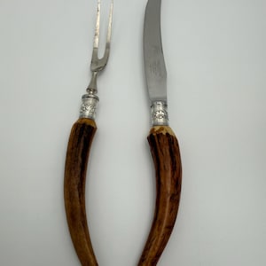May include: A two-piece carving set consisting of a two-tined fork and a curved knife. Both utensils feature natural brown textured antler handles with silver-toned metal ferrules decorated with floral engravings and stainless steel blades.