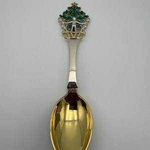 May include: A decorative metal spoon featuring a gold-toned bowl and a handle topped with a green enameled Christmas tree design. Two silver-toned figures stand at the base of the tree, and the year 1932 is engraved on the neck of the spoon.