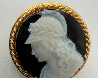 Könnte beinhalten: Runde Cameo-Brosche mit dem Profil einer Figur mit Helm und Lorbeerkranz, vor schwarzem Hintergrund. Die Figur ist weiß, und der Rahmen ist goldfarben mit einem seilartigen Rand.