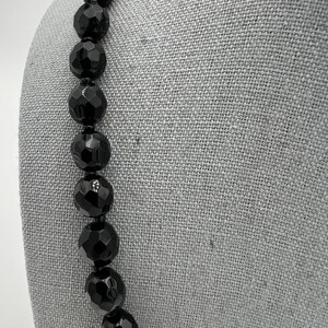 May include: A black beaded necklace with large, faceted beads. The beads are strung together on a black cord. The necklace is displayed on a gray surface.