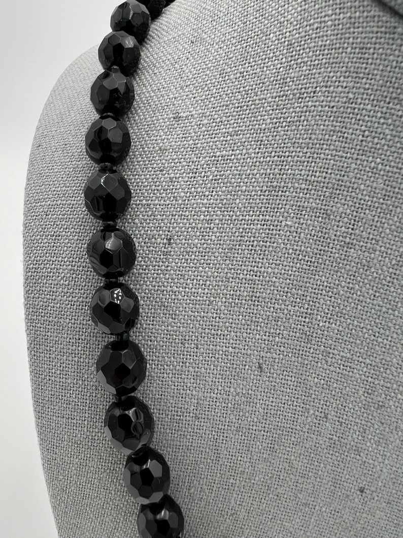 May include: A black beaded necklace with large, faceted beads. The beads are strung together on a black cord. The necklace is displayed on a gray surface.