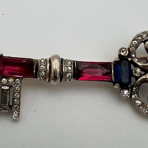 Op de afbeelding: Zilverkleurige sleutelvormige broche met een decoratief ontwerp. De sleutel heeft rechthoekige en ronde rode en blauwe edelstenen en heldere kristallen. De kop van de sleutel heeft een decoratief, opengewerkt ontwerp en de schacht van de sleutel is versierd met edelstenen.