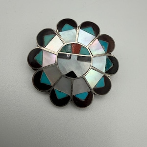 May include: A flower-shaped brooch with a silver-tone metal frame. The brooch features a central design of black, white, and red elements, surrounded by turquoise, white, and brown segments. The design is reminiscent of Native American artistry.