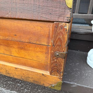 Puede incluir: Un ba&uacute;l de madera vintage con protectores de esquina de lat&oacute;n y un pestillo de metal. El ba&uacute;l tiene un tono de madera rico y c&aacute;lido con vetas visibles y probablemente se utiliza para almacenamiento o decoraci&oacute;n. El ba&uacute;l es un mueble cl&aacute;sico.