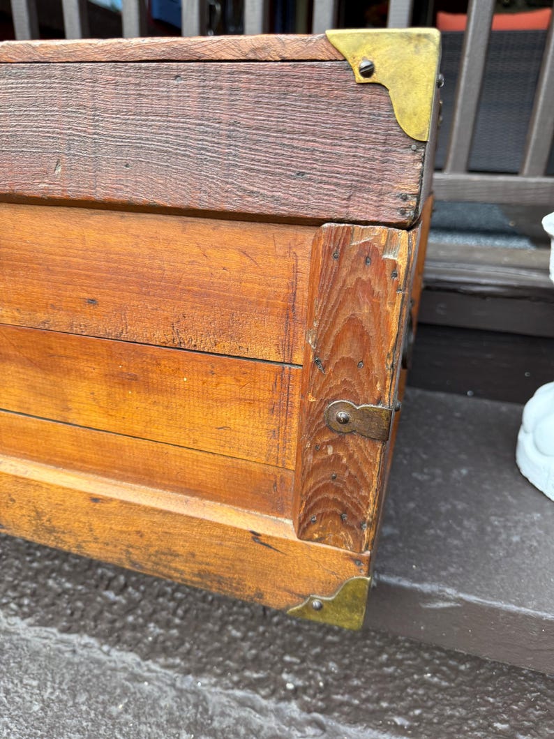 Puede incluir: Un ba&uacute;l de madera vintage con protectores de esquina de lat&oacute;n y un pestillo de metal. El ba&uacute;l tiene un tono de madera rico y c&aacute;lido con vetas visibles y probablemente se utiliza para almacenamiento o decoraci&oacute;n. El ba&uacute;l es un mueble cl&aacute;sico.