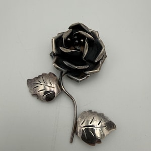 May include: A silver-toned rose brooch with detailed petals and two leaves. The brooch is a realistic depiction of a rose, with a dark center and textured leaves. The stem curves gently, connecting the flower and leaves.