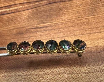 May include: A vintage gold-toned barrette featuring six oval-shaped, iridescent gemstones. The gemstones display a range of colors, including purple, green, and gold. The barrette is set against a wooden background.