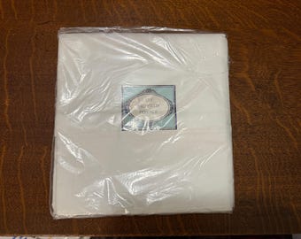 May include: A folded, white textile product, possibly bedding or linens, is encased in clear plastic. A label with the text "LADY SHEFFIELD PERCALE" is visible on the packaging. The product is on a dark wood surface.