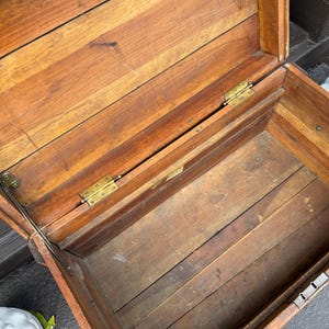 Puede incluir: Ba&uacute;l de madera abierto con tapa con bisagras, que revela un interior vac&iacute;o. El ba&uacute;l est&aacute; construido con tablones de madera, con vetas visibles y un c&aacute;lido tono marr&oacute;n rojizo. Bisagras y herrajes de lat&oacute;n visibles.