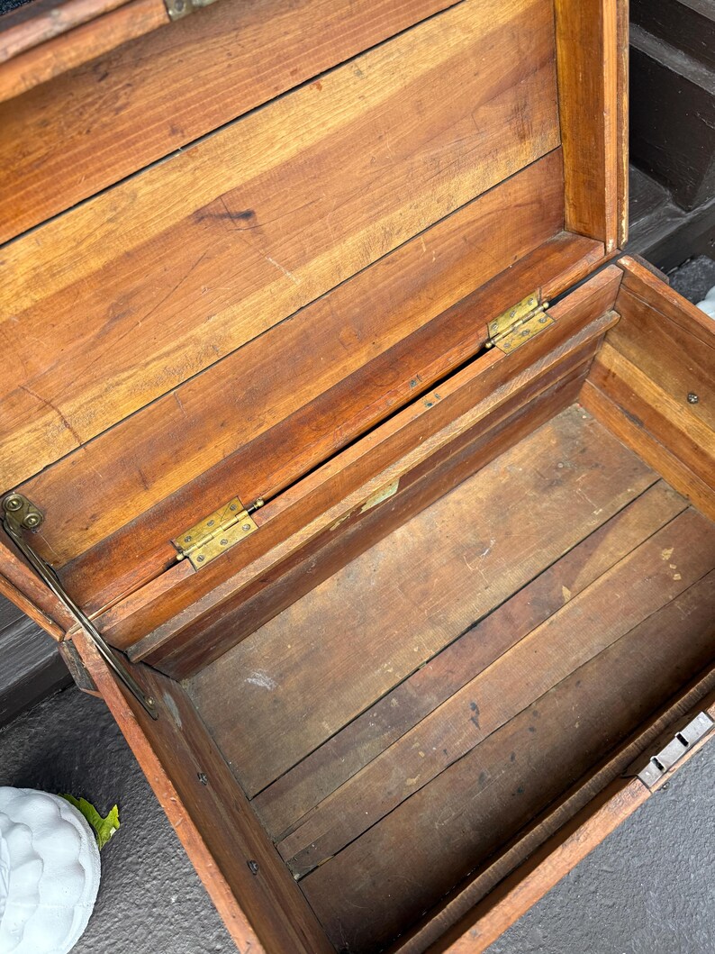 Puede incluir: Ba&uacute;l de madera abierto con tapa con bisagras, que revela un interior vac&iacute;o. El ba&uacute;l est&aacute; construido con tablones de madera, con vetas visibles y un c&aacute;lido tono marr&oacute;n rojizo. Bisagras y herrajes de lat&oacute;n visibles.